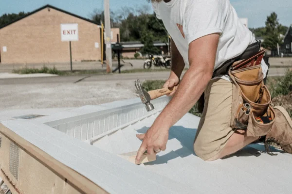 Bearded worker in cap and tool belt hammers metal roofing panel on a sunny day at a construction site.