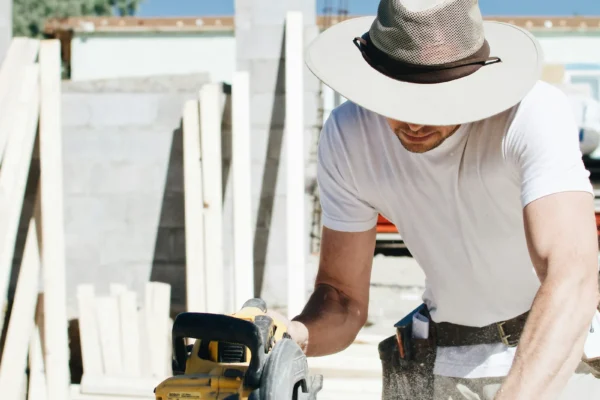 Construction worker in white hat and shirt using a circular saw on a sunny rooftop job site.