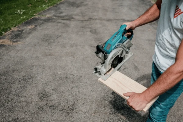 Man using a turquoise circular saw to cut a piece of light wood on a concrete driveway.