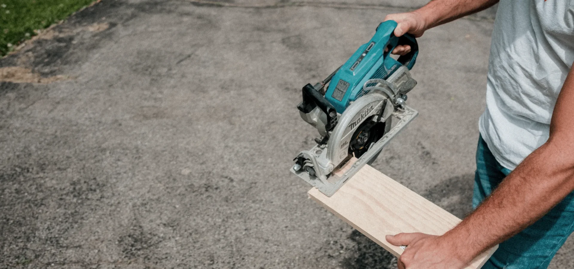 Man using a turquoise circular saw to cut a piece of light wood on a concrete driveway.