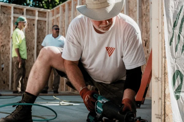 Worker in hat and gloves uses power tool on concrete floor in outdoor construction area with wooden framing.