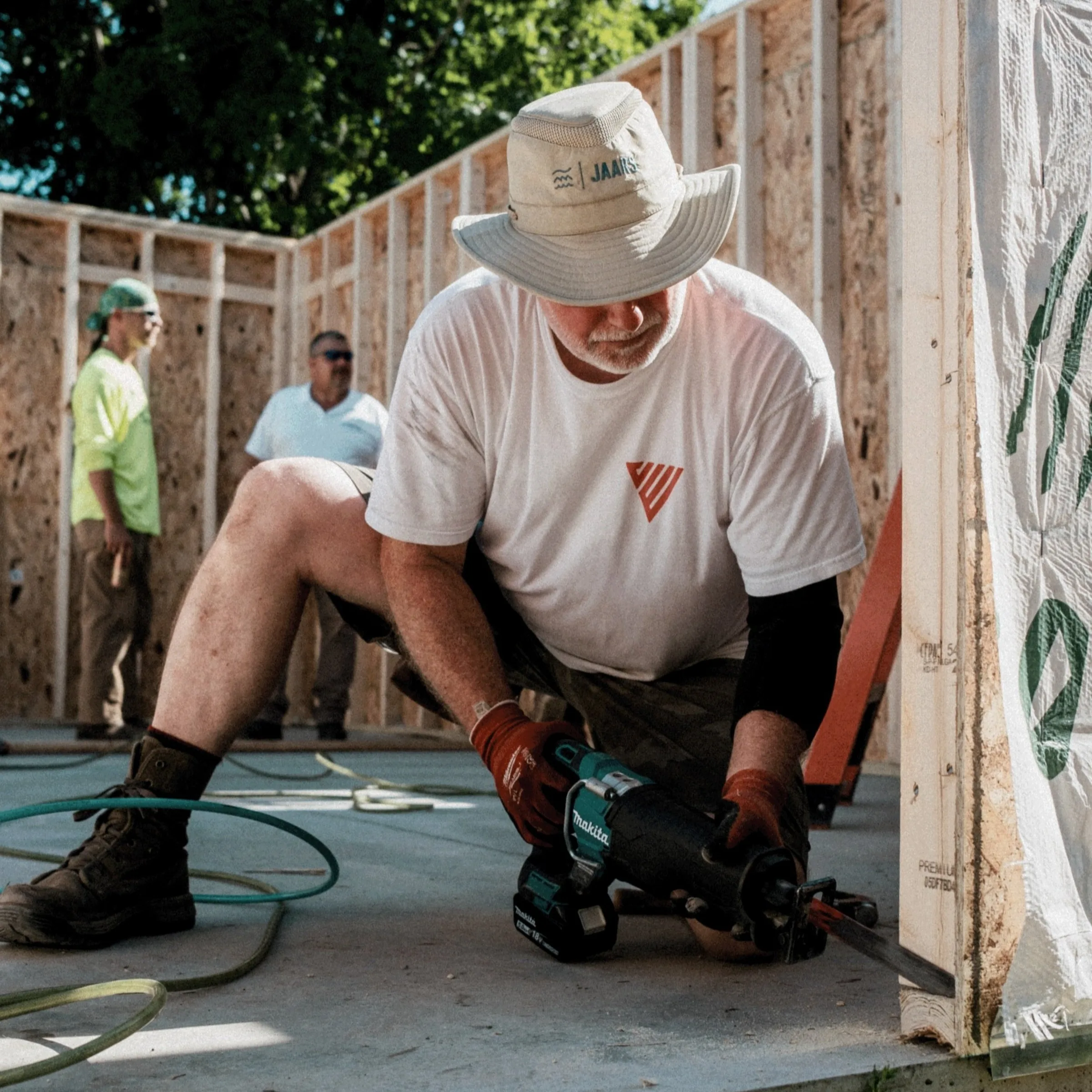 Worker in hat and gloves uses power tool on concrete floor in outdoor construction area with wooden framing.