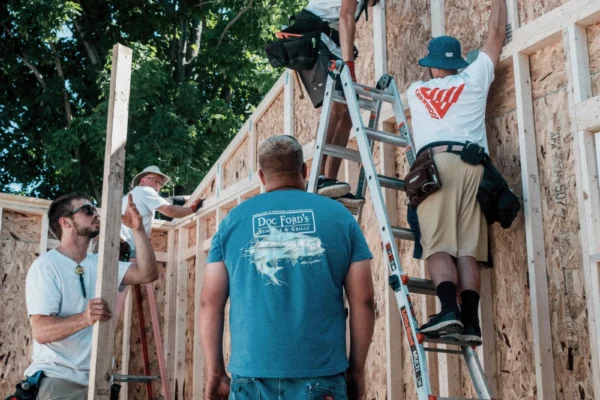 Construction workers build a wooden frame structure outdoors, with two men on ladders installing OSB panels while others assist below.
